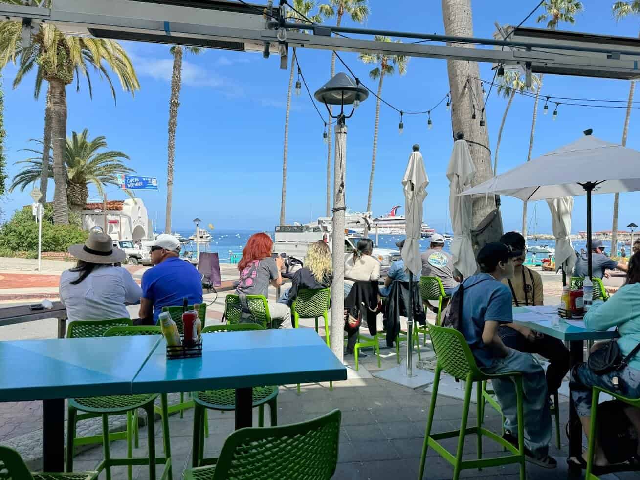 Outdoor seating at a casual café in Avalon, Catalina Island, where people are enjoying meals with an ocean view and a docked cruise ship in the distance.

