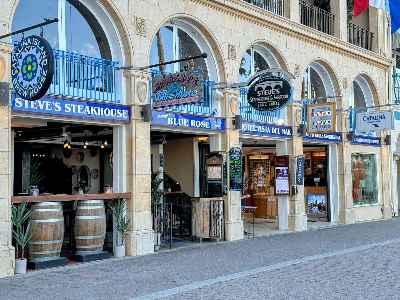 A row of Avalon storefronts on Catalina Island, including Steve’s Steakhouse and Maggie’s Blue Rose, with ornate architecture and hanging signs above each business.

