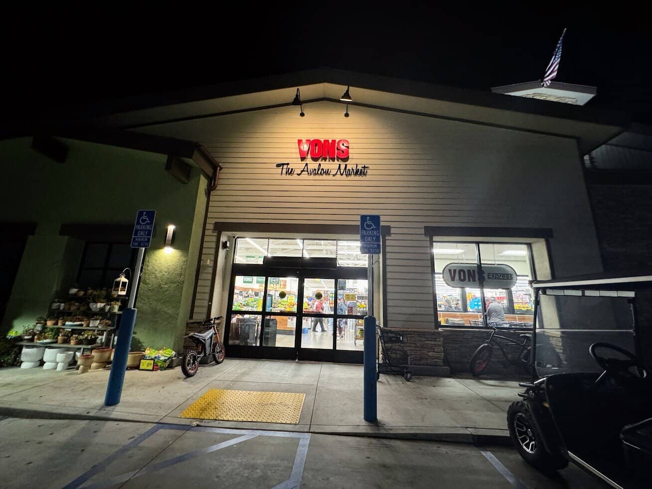Exterior of the Vons grocery store in Avalon, Catalina Island, lit up at night with customers entering, bikes and golf carts parked out front.

