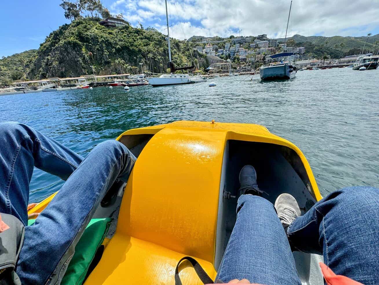 A view from a bright yellow paddle boat on the water, with two people’s legs visible, looking toward Catalina Island’s rugged shore lined with boats and colorful homes.

