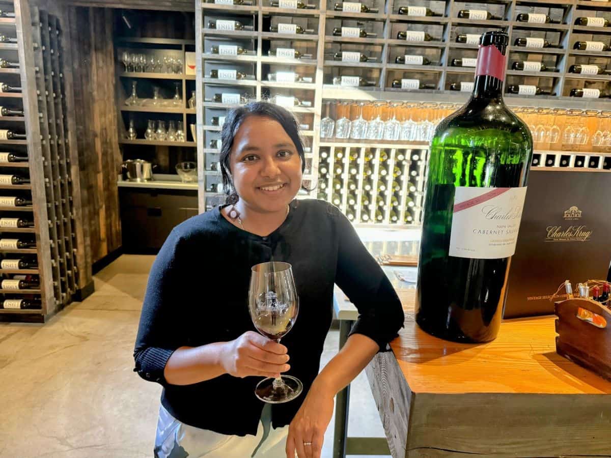 A smiling woman holding a wine glass poses beside an oversized bottle of Charles Krug Cabernet Sauvignon inside the historic wine tasting room, surrounded by shelves of vintage wine bottles—an iconic stop among the best things to do in St. Helena.