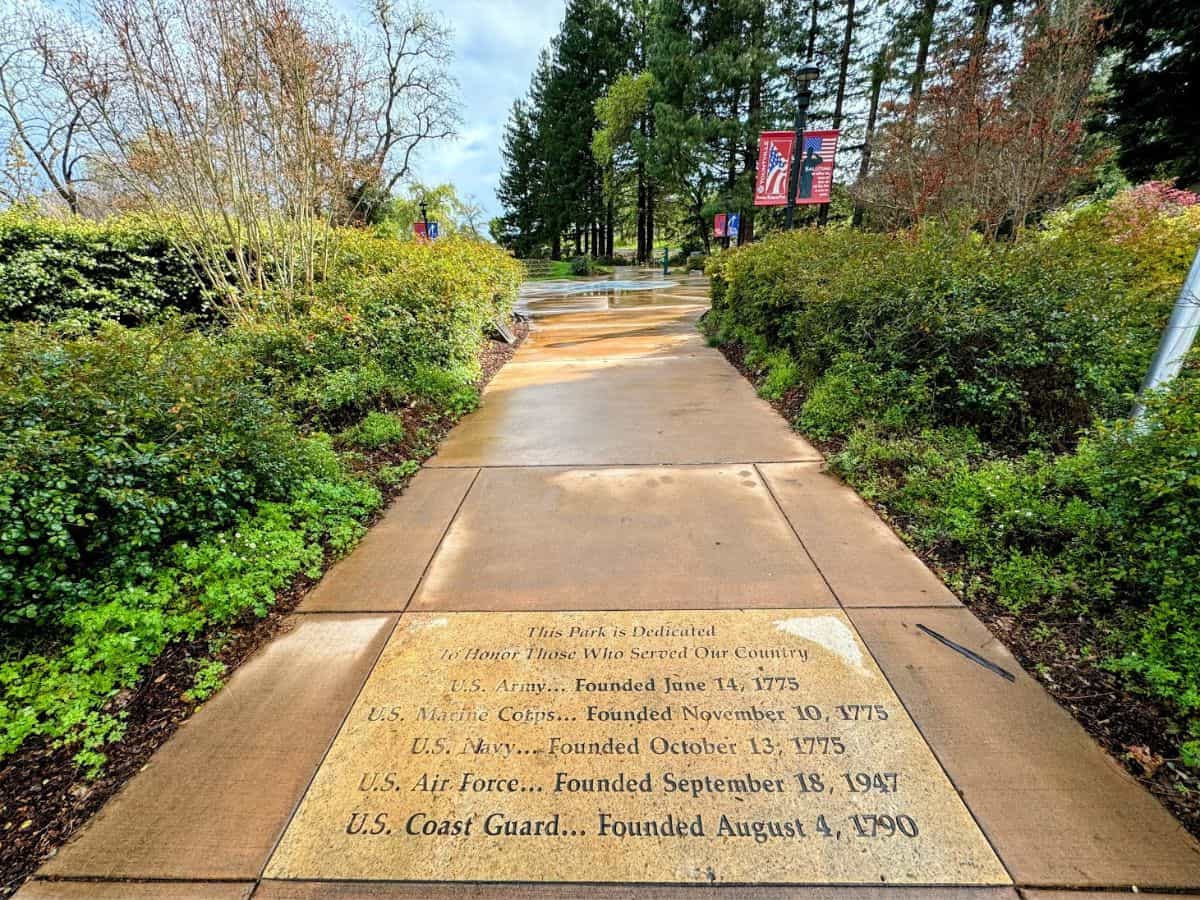 A wet walkway leads through a lush garden park with engraved stone honoring military branches, flanked by trees and American flags. Reflecting at Veterans Memorial Park is a meaningful and quiet thing to do in Yountville.