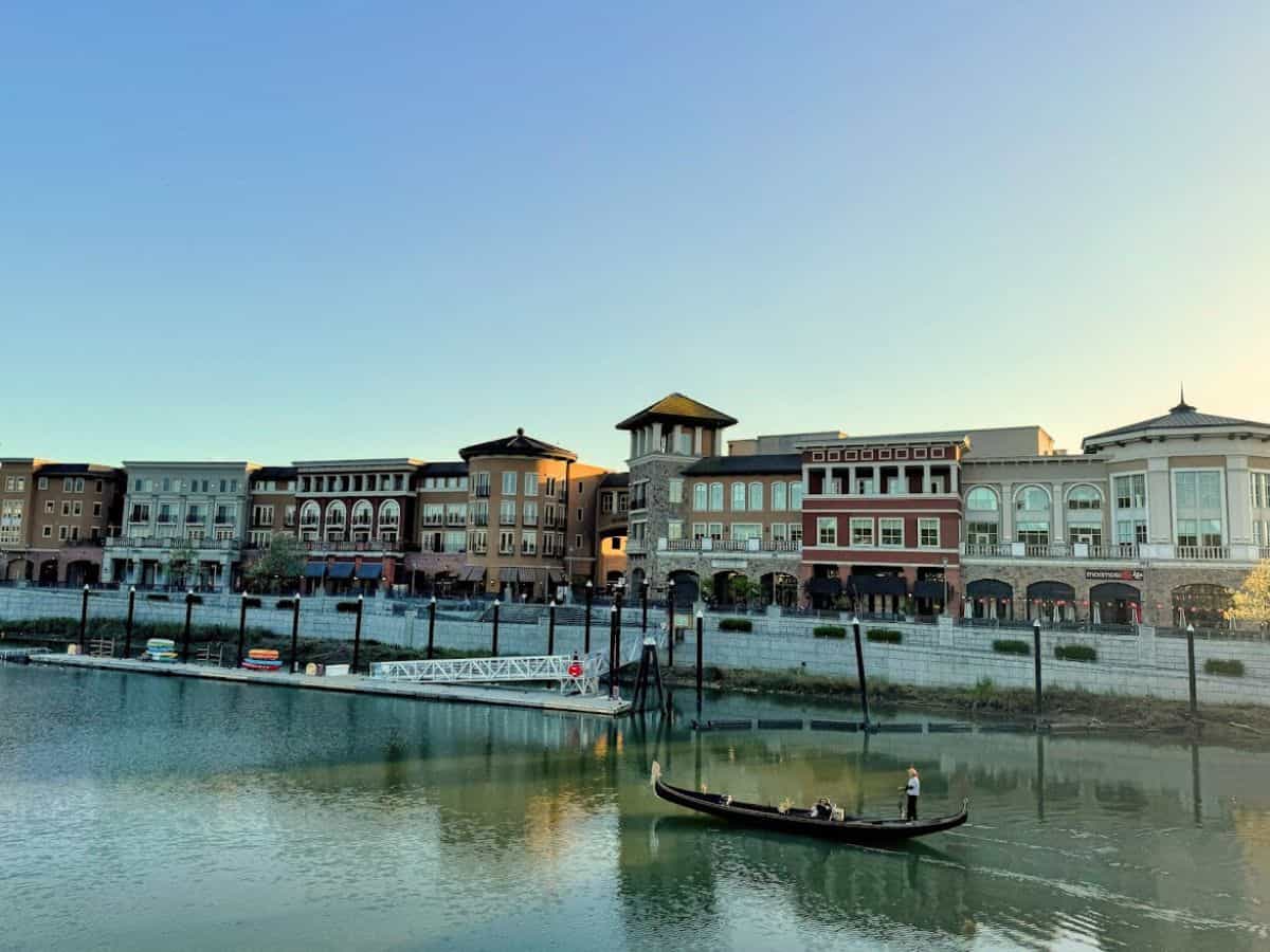 A gondolier steering a sleek black gondola down the Napa River with European-style buildings in the background—offering a romantic, Venetian-style experience on the list of 17 things to do in Napa.
