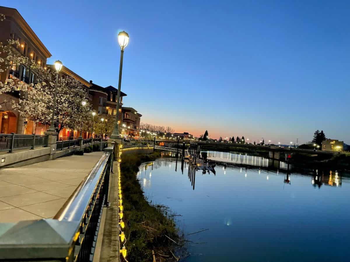 Scenic twilight view of the Napa Riverfront with glowing streetlights, cherry blossoms, and a calm river—ideal for a peaceful stroll on your list of 17 things to do in Napa.
