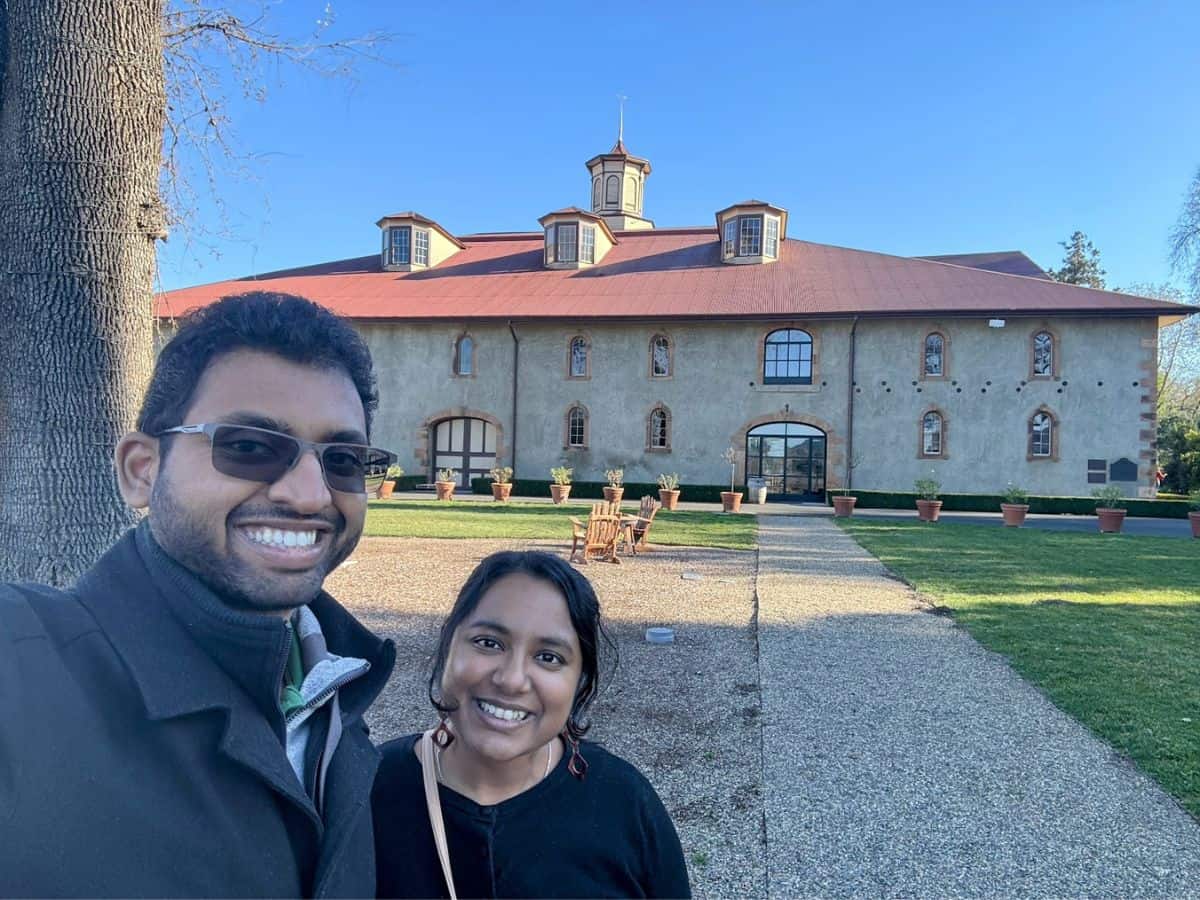 A smiling couple takes a selfie in front of the historic Charles Krug Winery building under clear blue skies, surrounded by a manicured lawn and terracotta potted plants—capturing one of the top things to do in St. Helena.