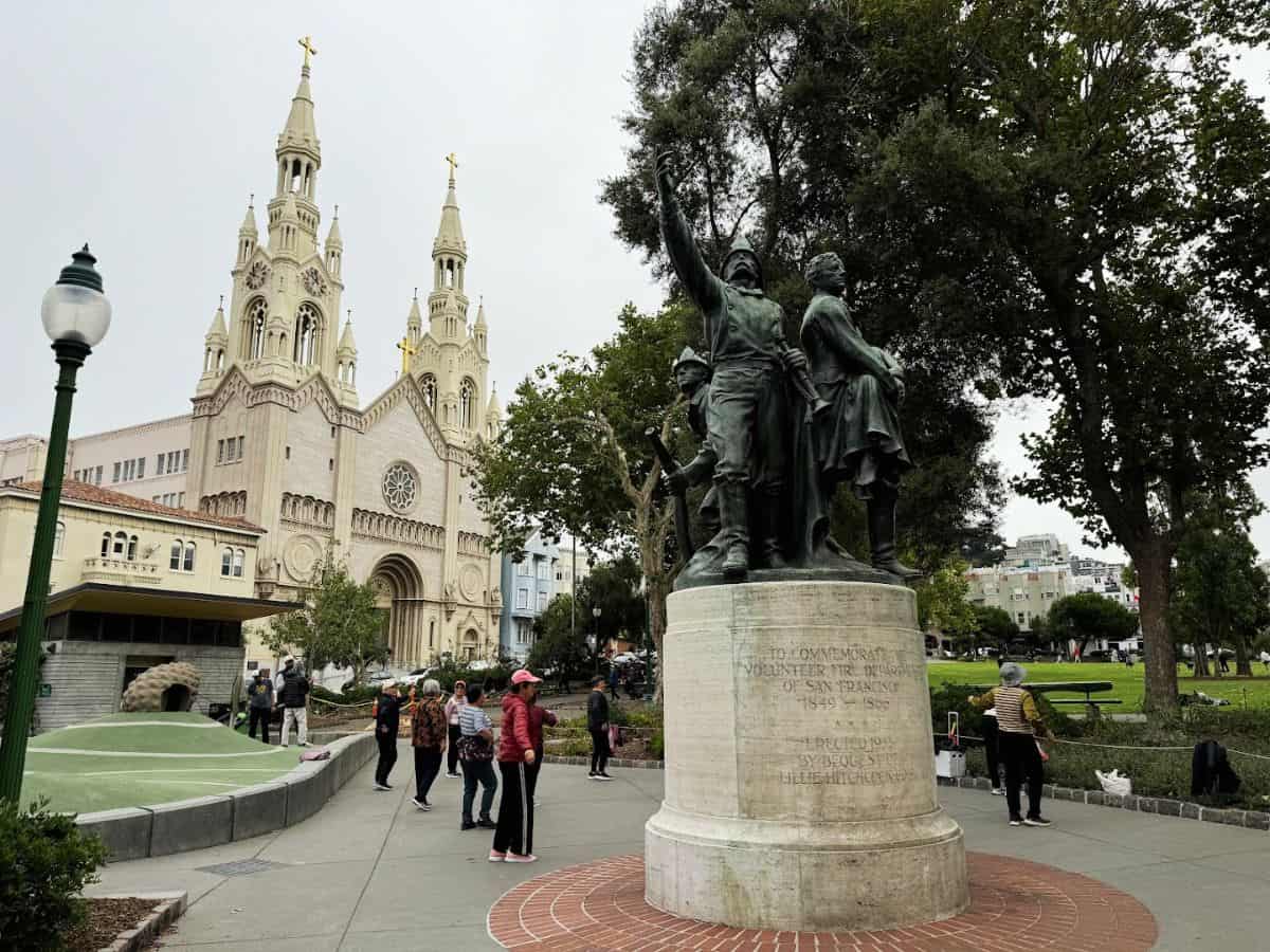Historic statue commemorating San Francisco's contributions, with iconic church and city park backdrop.