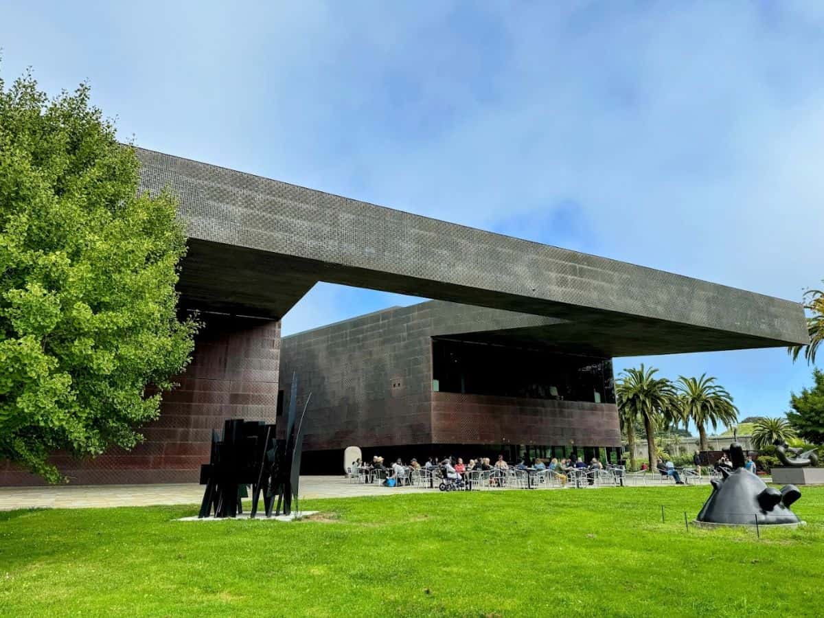 Modern contemporary building with outdoor seating in a park setting, featuring lush green grass, sculptures, and palm trees.