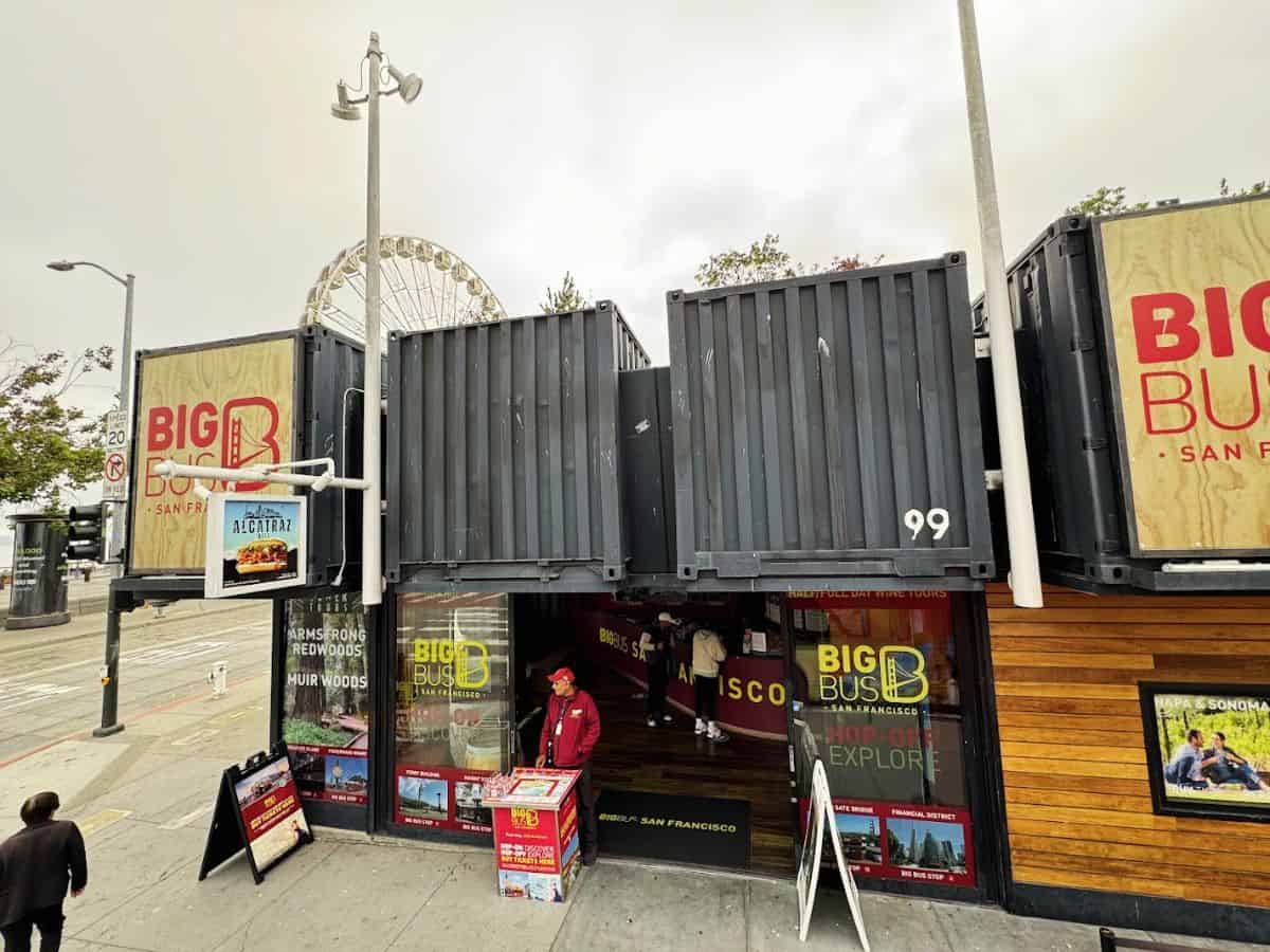 Big Bus Tour office at Fisherman’s Wharf with signs for hop-on hop-off tours and a ferris wheel peeking in the background