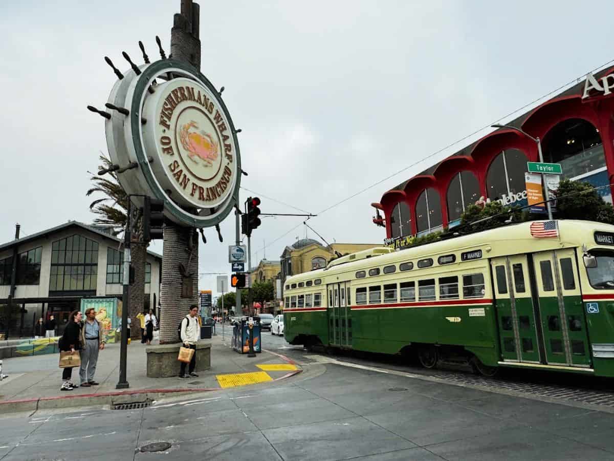 Famous Fisherman’s Wharf sign shaped like a ship wheel with a vintage green streetcar passing by on a cloudy day