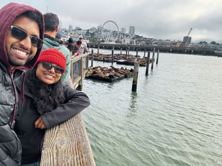 Smiling couple in jackets at Fisherman’s Wharf with sea lions resting on floating docks and the San Francisco skyline in the background