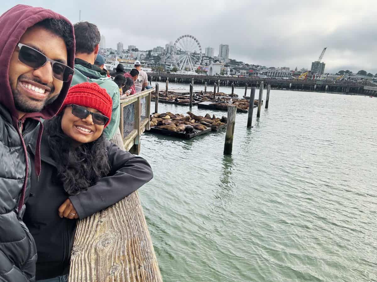Smiling couple in jackets at Fisherman’s Wharf with sea lions resting on floating docks and the San Francisco skyline in the background